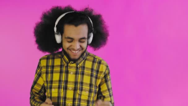 Un joven con un peinado africano sobre un fondo rosa está escuchando música y auriculares. Sobre un fondo de color — Vídeos de Stock
