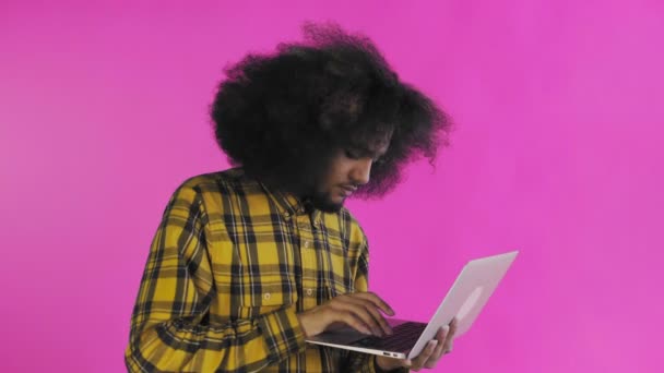 Молодий чоловік з африканською зачіскою на рожевому фоні друкує на ноутбуці. На кольоровому фоні — стокове відео