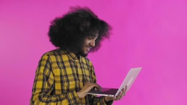 En ung mand med en afrikansk frisure på en lyserød baggrund skriver på en bærbar computer. På en farvet baggrund – Stock-video