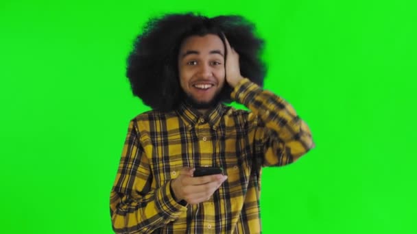 Um jovem com um penteado africano em um fundo verde olha para o telefone e fica feliz surpreso. Emoções em um fundo colorido — Vídeo de Stock