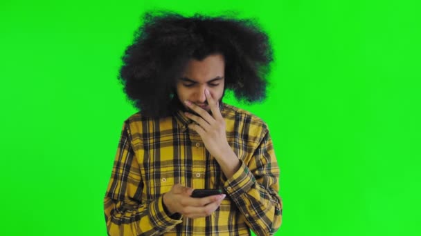 Ένας νεαρός άνδρας με αφρικανικό χτένισμα σε πράσινο φόντο κοιτάζει το τηλέφωνο και είναι ευτυχής έκπληξη. Συναισθήματα σε έγχρωμο φόντο — Αρχείο Βίντεο
