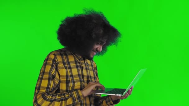 Un joven con un peinado africano sobre un fondo verde está escribiendo en un portátil. Sobre un fondo de color — Vídeos de Stock