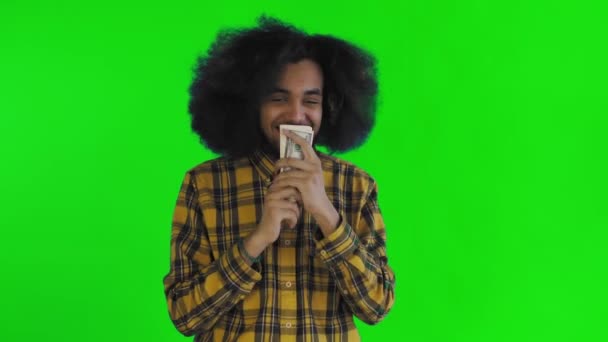 Szczęśliwy Afroamerykanin trzymający banknoty w rękach, całujący je i patrzący w kamerę, stoi odizolowany na zielonym tle. — Wideo stockowe