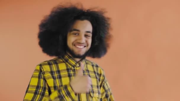 Un afro-americano feliz mirando a la cámara muestra un parecido con su mano, se encuentra aislado sobre un fondo naranja — Vídeos de Stock