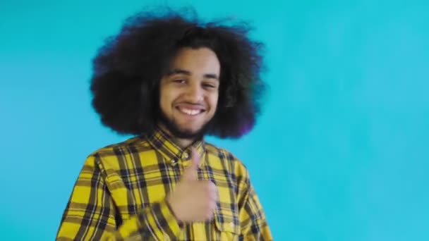 Un hombre afroamericano feliz mirando a la cámara, muestra un parecido con su mano, se encuentra aislado sobre un fondo azul — Vídeos de Stock