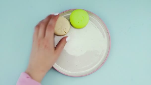 Κάτοψη των πολύχρωμων γαλλικών μπισκότων Macarons σε παστέλ μπλε φόντο. Ένα γυναικείο χέρι βάζει μπισκότα σε ένα πιάτο — Αρχείο Βίντεο