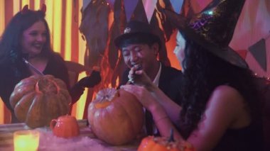 İki kız ve ürkütücü Cadılar Bayramı kostümlü bir adam bir masada oturmuş sohbet ediyor ve balkabaklarına yüzler oyuyorlar.