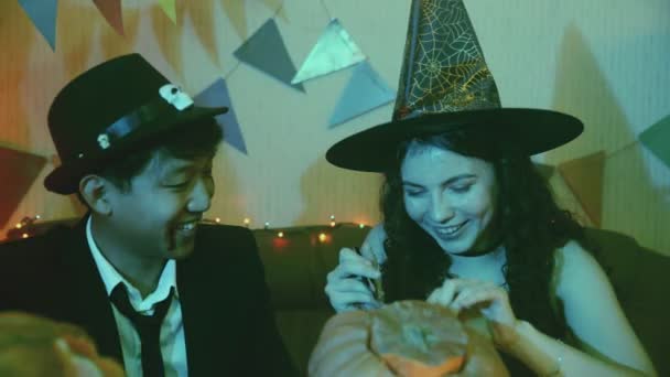 Dívka a chlap v děsivých halloweenských kostýmech sedí u stolu a vyřezávají tváře na dýni — Stock video