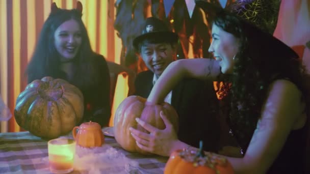 Twee meisjes en een man in griezelige Halloween kostuums zitten aan een tafel en halen de pulp uit pompoenen — Stockvideo