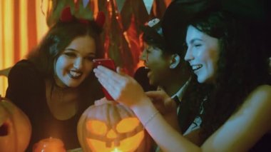 Bir grup arkadaş ürkütücü kostümlerle bir masada oturuyor ve Cadılar Bayramı 'nda telefona bakıyor ve gülüyorlar.