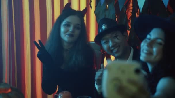 Группа друзей сидит за столом в жутких костюмах, фотографируется с камерой и смеется в Хэллоуин — стоковое видео