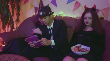Ürkütücü kostümlü bir adam tabletle oynuyor ve kızlar Cadılar Bayramı partisinde telefonla konuşuyorlar..