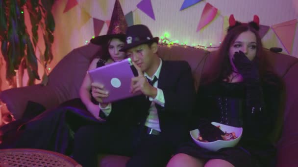 Een man in een griezelig kostuum speelt actief op een tablet, en de meisjes zitten en vervelen zich op een Halloween feest. — Stockvideo