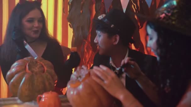 Две девушки и парень в жутких костюмах на Хэллоуин сидят за столом и болтают и вырезают лица на тыквах — стоковое видео