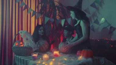 İki kız ve ürkütücü Cadılar Bayramı kostümü giymiş bir adam bir masada oturmuş balkabaklarının posasını çıkarıyorlar.