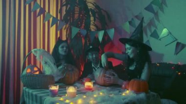 İki kız ve ürkütücü Cadılar Bayramı kostümü giymiş bir adam bir masada oturmuş balkabaklarının posasını çıkarıyorlar.