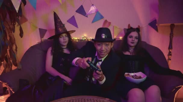 Chlápek v děsivém kostýmu aktivně hraje videoherní konzoli a holky sedí a nudí se na Halloweenské párty. — Stock video