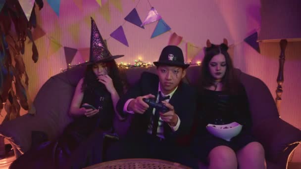 El tipo con el disfraz espeluznante está jugando activamente a la consola, y las chicas están sentadas en el teléfono en la fiesta de Halloween. — Vídeos de Stock