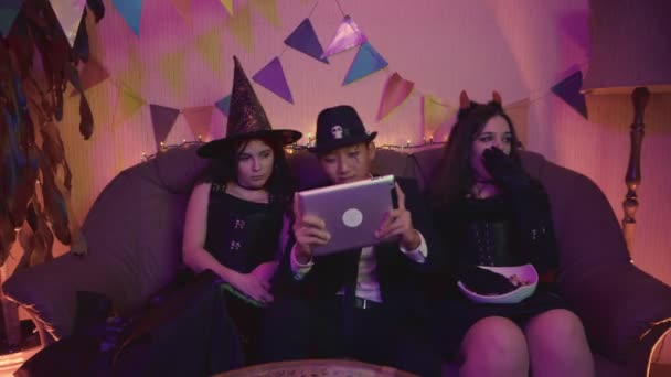 Een man in een griezelig kostuum speelt actief op een tablet, en de meisjes zitten en vervelen zich op een Halloween feest. — Stockvideo