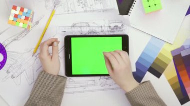 Üst manzara. Tasarımcı bir kız krom ekranda tabletle masada oturuyor. Masada çeşitli ofis malzemeleri var.