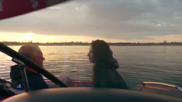 En kille och en tjej sitter i en motorbåt och kysser och tittar på solnedgången. Romantisk atmosfär. — Stockvideo