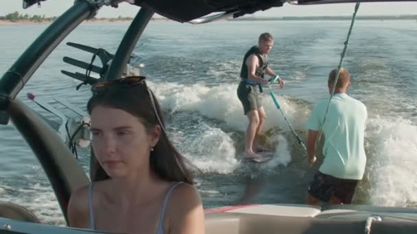 Una mujer está conduciendo un barco a motor, y en el fondo un tipo está montando un wakeboard, y el hombre lo está ayudando — Vídeo de stock
