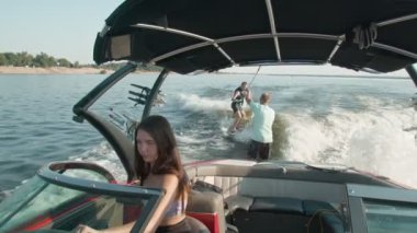 Bir kadın motorlu tekne kullanıyor, ve arka planda bir adam wakeboard kullanıyor, ve erkek ona yardım ediyor.
