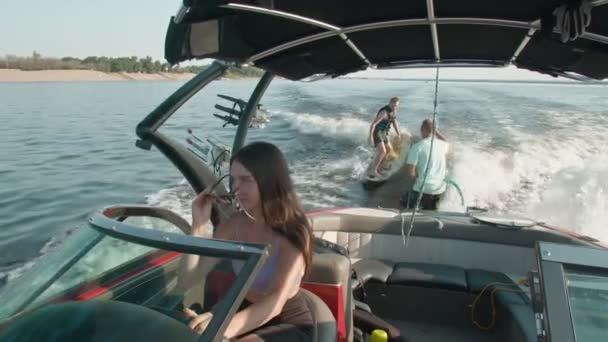 Uma mulher está dirigindo um barco a motor, e no fundo um cara está montando um wakeboard, e o homem está ajudando-o — Vídeo de Stock