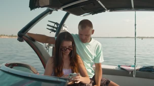 En man och en kvinna sitter i en motorbåt och tittar på telefonen och pratar. — Stockvideo