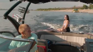 Bir adam motorlu tekne kullanıyor, ve arka planda bir kadın oturuyor ve hoş havanın tadını çıkarıyor.