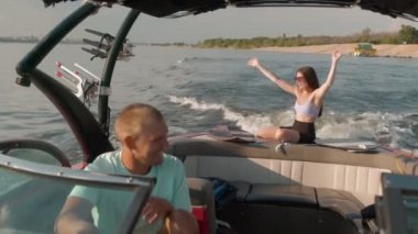 Bir adam motorlu tekne kullanıyor, ve arka planda bir kadın oturuyor ve hoş havanın tadını çıkarıyor.