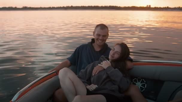 Um cara e uma menina abraçar bonito ao pôr do sol sentado em um barco — Vídeo de Stock