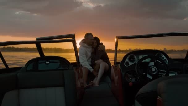 Facet i dziewczyna siedzą na motorówce, całując się i oglądając zachód słońca. Romantyczna atmosfera. — Wideo stockowe