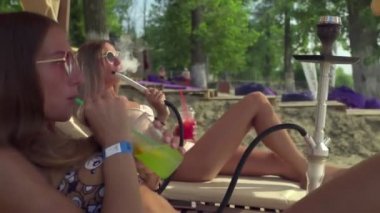 Kumsalda güneşlenen iki genç kadın nargile içiyor ve kokteyl içiyor.