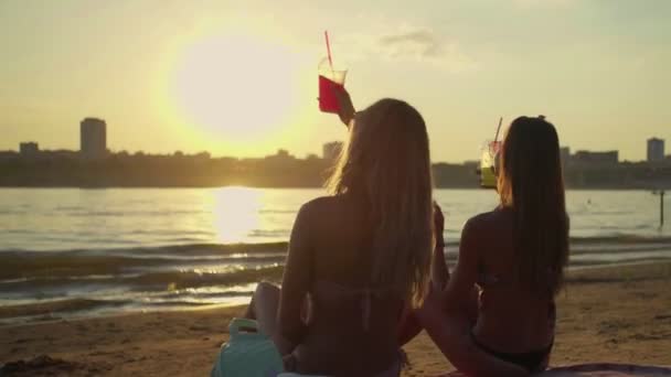 Две счастливые девушки на пляже пьют коктейли и смотрят на закат — стоковое видео