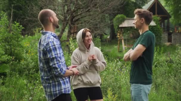 Um grupo de três pessoas está falando e rindo enquanto está em pé em uma floresta cercada por árvores em um dia de verão. Ao ar livre — Vídeo de Stock