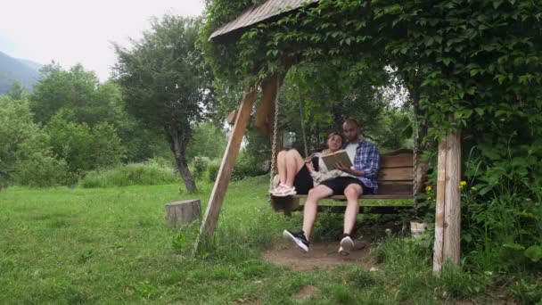 Bir adam ve bir kadın ahşap salıncakta oturuyorlar. Bir adam bir kızla kitap okuyor. Bir erkekle bir kızın randevusu var. Doğada — Stok video