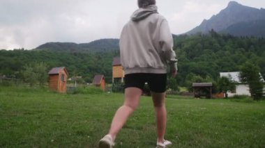 Dişi bir turist dağların ve ahşap evlerin arasında çimenlerde yürüyor. Doğa ve dağlar