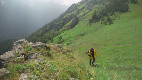 Na horu se vyšplhá turista ve žlutém plášti do deště s velkým batohem na zádech. Cestovní ruch. — Stock video
