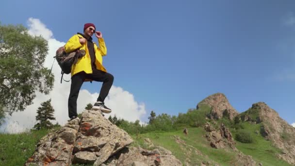 En manlig turist sätter sin fot på en klippa och tittar in i fjärran i en gul regnrock, med en stor ryggsäck på ryggen. Turism. — Stockvideo