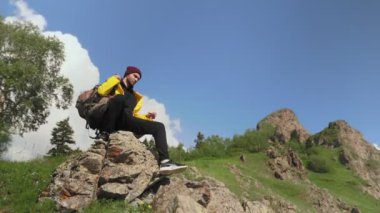 Sırtında büyük bir sırt çantası olan sarı yağmurluk giymiş bir erkek turist bir kayanın üzerinde oturuyor ve doğanın tadını çıkarıyor. Turizm.