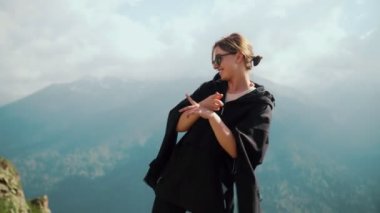 Güneş gözlüklü ve siyah kapüşonlu mutlu bir genç kız dağların arkasında dans ediyor.