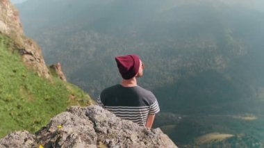 Sırtı dağların arka planında, bir taşın üzerinde oturan şapkalı genç bir adamın arka görüntüsü.