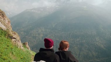 Genç bir adam ve bir kız dağlık bir bölgede bir kayanın üzerinde oturuyorlar ve güzel manzaralara bakıyorlar. Turizm