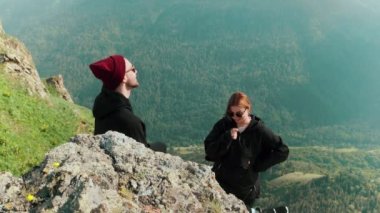 Genç bir adam ve bir kız dağlık bir bölgede bir kayanın üzerinde iletişim kuruyor ve güzel manzaralara bakıyor. Turizm