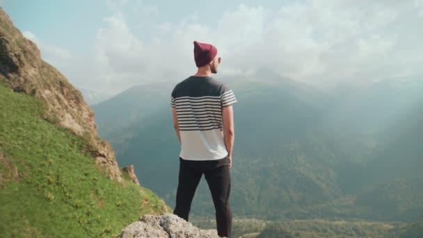 Visão traseira de um jovem com um chapéu de pé com as costas no fundo das montanhas — Vídeo de Stock