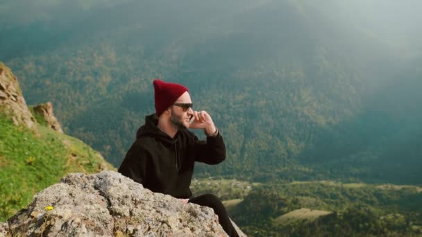 En ung resenär sitter på klipporna använder sin telefon för att chatta med människor på nätet och dela hisnande landskap i bakgrunden. — Stockvideo