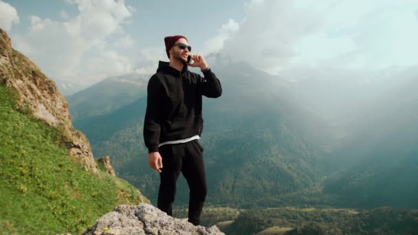 Een jonge reiziger, die in de bergen staat, gebruikt zijn telefoon om online met mensen te chatten en het adembenemende landschap op de achtergrond te delen. — Stockvideo