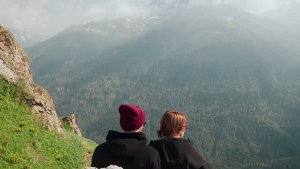 Genç bir adam ve bir kız dağlık bir bölgede bir kayanın üzerinde oturuyorlar ve güzel manzaralara bakıyorlar. Turizm — Stok video