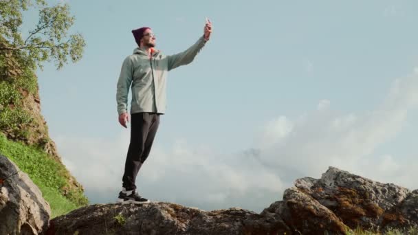 Mladý muž v klobouku a slunečních brýlích, stojí na skalách a bere si do telefonu selfie. Cestování — Stock video